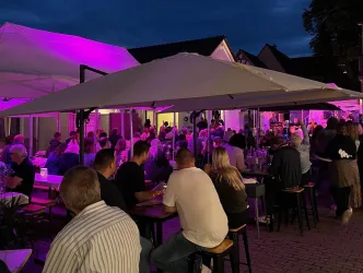 Ein belebtes Restaurant mit vielen Gästen unter Sonnenschirmen. Die Umgebung ist Abenddämmerung mit bunten Lichtern.