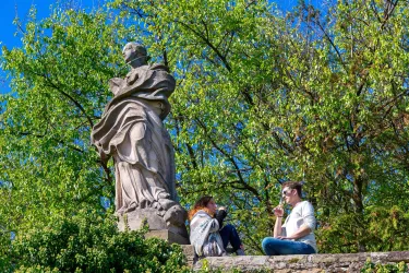 Zwei Personen sitzen neben einer Statue in einem Garten. Der Himmel ist blau und die Bäume sind grün.