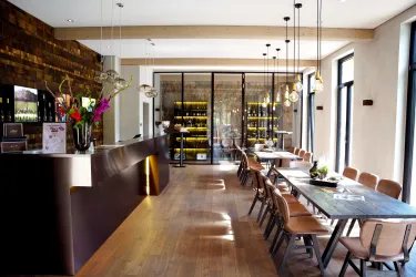 Ein modernes Restaurant mit einem eleganten Empfangsbereich und langen Holztischen. Große Fenster lassen viel Licht herein und sorgen für eine einladende Atmosphäre.