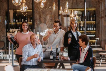Eine Gruppe von fünf Personen steht in einem eleganten Raum mit Wein. Sie lächeln und halten Gläser, während im Hintergrund Regale mit Weinflaschen zu sehen sind.