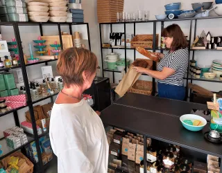 In einem Geschäft stehen zwei Frauen. Eine Verkäuferin packt Produkte in Papier, während die Kundin zusieht.