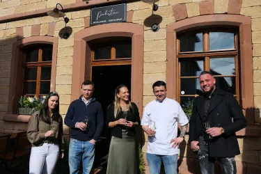 Eine Gruppe von fünf Personen steht lächelnd vor einem Restaurant. Sie halten Gläser in der Hand und posieren vor einer steinernen Wand mit Fenstern.