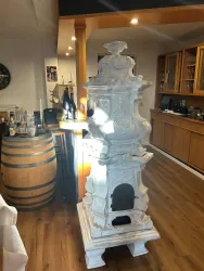 Ein eleganter Kachelofen steht in einem gemütlichen Raum. Im Hintergrund sind Holzregale und ein Fass zu sehen.