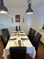 Ein schön gedeckter Esstisch mit schwarzen Stühlen und einer weißen Tischdecke. An der Wand hängt ein Bild und die Beleuchtung wird von zwei Pendelleuchten erzeugt.