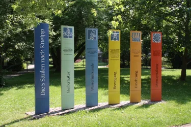Sechs farbige Säulen stehen in einem Park und repräsentieren die fünf Säulen nach Kneipp. Sie sind umgeben von grünem Grass und Bäumen.