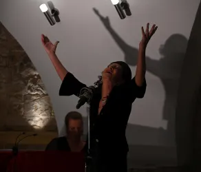 Eine Sängerin steht auf der Bühne und hebt ihre Arme ausdrucksvoll. Im Hintergrund ist ein Musiker am Klavier sichtbar.