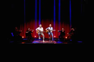 Zwei Musiker spielen live auf der Bühne Gitarre und singen. Im Hintergrund stehen weitere Musiker mit Streichinstrumenten.