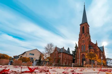 Eine beeindruckende Kirche mit einem hohen Turm und roten Blättern im Vordergrund. Der Himmel ist blau mit leichten Wolken.