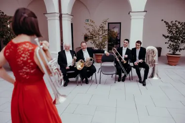 Eine Musikerin in einem roten Kleid spielt vor einer Band. Im Hintergrund sitzen vier Musiker mit verschiedenen Blasinstrumenten.