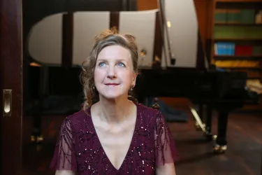 Eine Frau mit einem freundlichen Ausdruck sitzt vor einem Klavier. Der Raum hat eine elegante Atmosphäre mit Bücherregalen im Hintergrund.