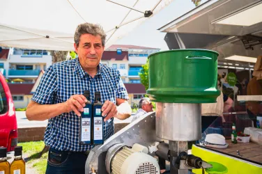 Ein Mann steht an einem Stand und hält eine Flasche in der Hand. Im Hintergrund sind weitere Produkte und ein grüner Behälter zu sehen.