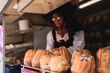 Eine Verkäuferin in traditioneller Kleidung steht an einem Verkaufsstand. Vor ihr liegen frisch gebackene Brötchen in Folie verpackt.