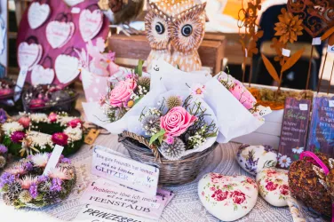 Ein schöner Korb mit bunten Blumen und einer rosa Rose steht auf einem Tisch. Im Hintergrund sind verschiedene Dekorationsartikel und Herzen zu sehen.