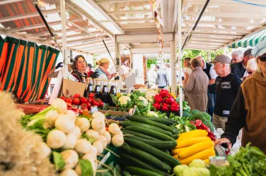 Ein belebter Markt mit frischem Gemüse und Obst. Menschen stehen in der Nähe der Verkaufsstände und genießen die Atmosphäre.