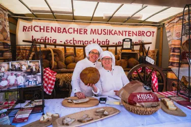 Zwei Bäckerinnen in weißen Schürzen präsentieren frisch gebackenes Brot an einem Marktstand. Im Hintergrund sind weitere Brote und eine bunte Verkaufsdekoration zu sehen.