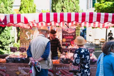 Ein Marktstand mit rot-weißem Dach, der verschiedene Wurstwaren anbietet. Kunden betrachten die Auswahl und interagieren mit dem Verkäufer.