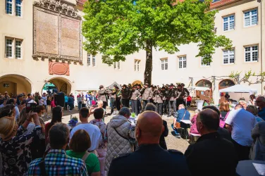 Eine lebhafte Veranstaltung in einem Innenhof mit vielen Menschen. In der Mitte steht eine Gruppe von Künstlern, die eine Aufführung zeigen.