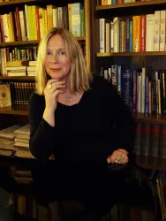 Eine Frau mit blonden Haaren sitzt an einem Tisch und blickt nachdenklich. Im Hintergrund sind viele Bücher in einem Regal zu sehen.