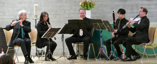 Eine Musikkapelle mit fünf Mitgliedern spielt auf einer Bühne. Sie sind in formeller Kleidung und verwenden verschiedene Blasinstrumente.