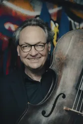 Ein lächelnder Mann hält ein Cello in den Händen. Im Hintergrund sind bunte Graffiti zu sehen.