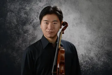 Ein junger Mann steht mit einer Violine in der Hand vor einem grauen Hintergrund. Er trägt ein schwarzes Hemd und hat einen neutralen Gesichtsausdruck.