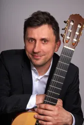 Ein Mann in einem schwarzen Anzug hält eine Gitarre. Er lächelt freundlich in die Kamera.