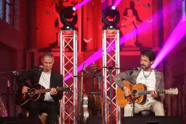 Zwei Musiker spielen akustische Gitarren auf einer Bühne. Bunte Lichter strahlen im Hintergrund und schaffen eine festliche Atmosphäre.