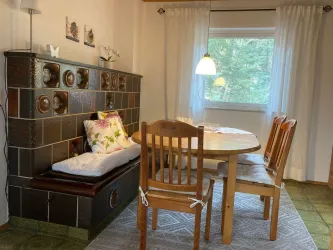 Ein gemütlicher Essbereich mit einem runden Tisch und vier Stühlen. Im Hintergrund ist ein Kachelofen und ein großes Fenster, das Tageslicht hereingleitet.