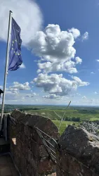 Eine Landschaft mit sanften Hügeln, einer Stadt im Hintergrund und einer Europaflagge vor einer beeindruckenden Wolkenformation. Der Himmel ist klar und sonnig.