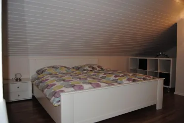 Ein gemütliches Schlafzimmer im Dachgeschoss mit einem weißen Bett und bunten Bettdecken. Neben dem Bett steht ein Nachttisch und es gibt ein Regal an der Wand.