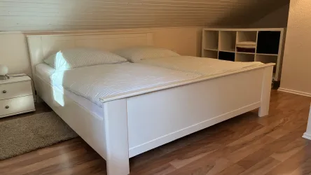 Ein gemütliches Schlafzimmer mit einem großen, weißen Doppelbett. Neben dem Bett steht ein Nachttisch und im Hintergrund ist ein Regal zu sehen.