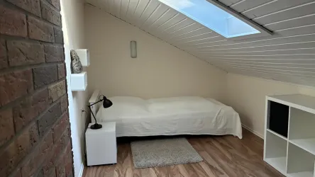 Ein gemütliches Zimmer mit einer schrägen Decke und einem Einzelbett. Es gibt einen Bücherregal und eine kleine Nachttischlampe.