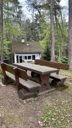 Ein rustikaler Tisch mit Bänken steht im Wald. Im Hintergrund ist unser Ferienhaus zwischen den Bäumen zu sehen.