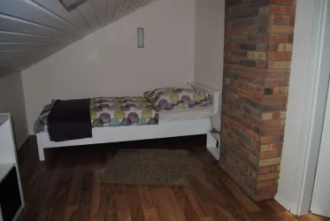 Ein einfaches Schlafzimmer mit einem Einzelbett und bunten Bettwäsche. Der Raum hat einen Holzboden und eine Steinwand.