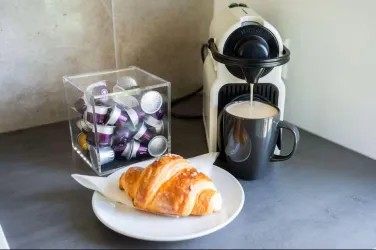 Eine Kaffeetasse steht neben einer Nespresso-Maschine, während ein Croissant auf einem weißen Teller serviert wird. Im Hintergrund befinden sich eine durchsichtige Box mit Kaffeekapseln.