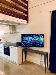 Ein modernes Wohnzimmer mit einer offenen Küche. Ein großer Fernseher steht auf einem schmalen Tisch, und im Hintergrund ist ein Schlafboden sichtbar.