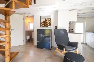 Ein gemütlicher Raum mit einer Wendeltreppe und einem modernen Sessel. Es gibt einen schlichten Schrank und eine offene Küche im Hintergrund.