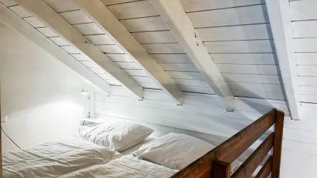 Ein gemütliches Schlafzimmer mit einem Doppelbett und einem Holzgeländer. Die Decke hat schräg angeordnete Holzbalken und die Wände sind hell gestrichen.