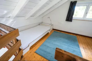Ein einfaches Schlafzimmer mit einem Einzelbett und einem blauen Teppich. Das Zimmer hat weiß getäfelte Wände und ein großes Fenster mit Vorhängen.