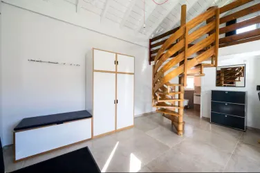Ein modernes, helles Zimmer mit einer Holztreppe und minimalistischem Mobiliar. Die Wände sind in neutralen Farben gehalten und der Boden ist aus hellem Stein.