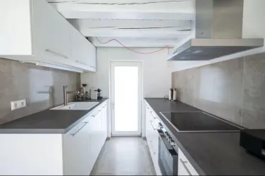 Eine moderne Küche mit weißen Schränken und einer grauen Arbeitsplatte. Das Licht fällt durch eine große Glastür, die nach draußen führt.