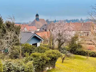 Eine ruhige Landschaft mit einem Blick auf ein Dorf, in dem eine Kirche mit einem Turm sichtbar ist. Umgeben von Bäumen und blühenden Pflanzen, bietet die Szene eine entspannte Atmosphäre.