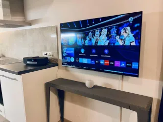Ein moderner Fernseher an einer Wand in einer Küche. Der Bildschirm zeigt eine Sendung mit mehreren Personen.