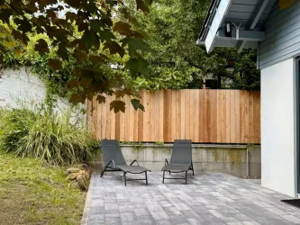 Ein ruhiger Außenbereich mit zwei Liegestühlen auf einer gepflasterten Fläche. Im Hintergrund steht ein Holzzaun und üppige Pflanzen sind zu sehen.
