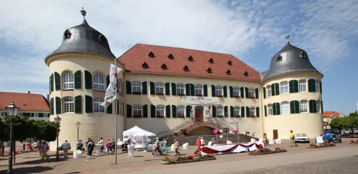 Ein historisches Gebäude mit runden Türmen und grünen Fensterläden. Vor dem Gebäude sind Menschen versammelt und Stände aufgebaut.