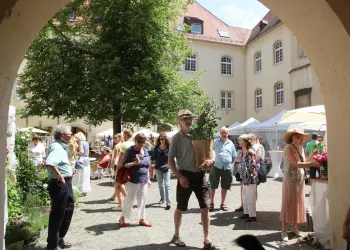 Ein belebter Markt mit vielen Menschen, die durch einen Innenhof schlendern. Stände mit Blumen und Pflanzen sind im Hintergrund sichtbar.