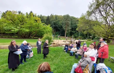 Eine Gruppe von Menschen sitzt im Freien auf einer Wiese. Im Vordergrund stehen einige Personen, die Informationen oder Anweisungen geben, während Bäume im Hintergrund sichtbar sind.