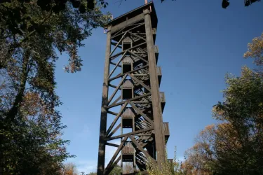 Ein großer Holzturm umgeben von Bäumen und blauem Himmel. Der Turm hat mehrere Stockwerke und Fenster.