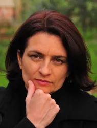 Eine nachdenkliche Frau mit kurzem, braunem Haar blickt direkt in die Kamera. Sie trägt einen schwarzen Mantel und hat eine Hand unter das Kinn gelegt.
