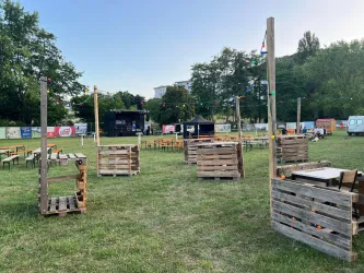 Eine Veranstaltung im Freien mit einfachen Holzmöbeln und einer Bühne im Hintergrund. Im Hintergrund sind viele Bäume und eine grüne Wiese sichtbar.